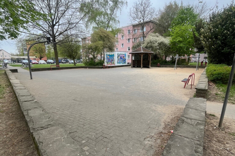 Spielplatz mit Basketballkorb, Schaukel und Pavillon, umgeben von Bäumen und Wohngebäuden. Ideal für Familien und Kinder.