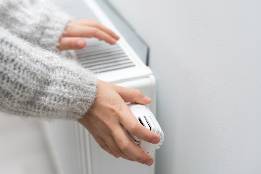 Hand, die den Thermostat eines Heizkörpers dreht, um die Temperatur zu regulieren. Weicher, grauer Pullover und helle Wand im Hintergrund.